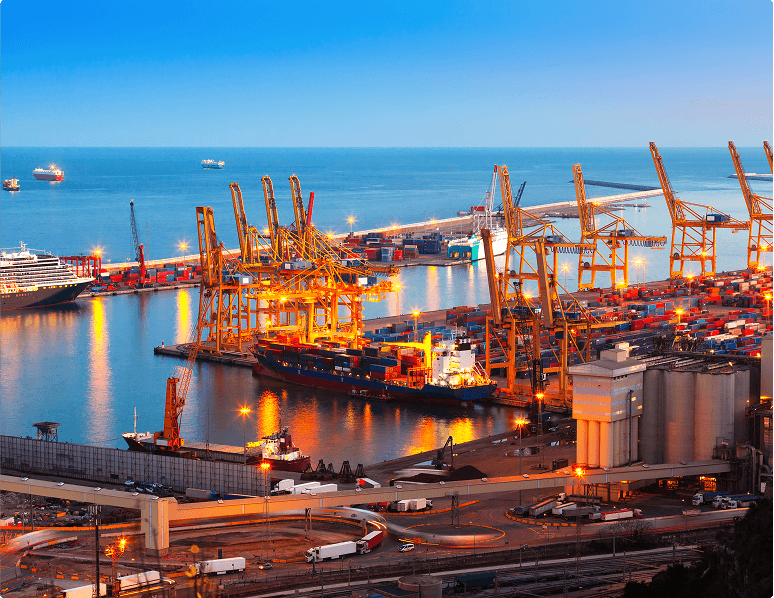 Vista de um porto à noite com guindastes iluminados, navios cargueiros e contêineres empilhados refletindo no mar.